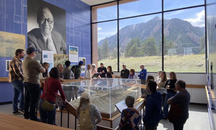 A group of people standing around the 3D model of the Mesa Lab during a tour. Behind them is a photo of I.M. Pei, the architect, and the Boulder flatiron mountains outside the window.